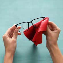 Eine Person hält eine Brille in der einen Hand und reinigt sie mit einem roten Microfaser Brillentuch, das in der anderen Hand gehalten wird. Der Hintergrund ist einfarbig türkis.