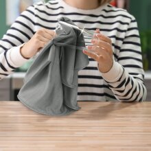 Person mit gestreiftem Pullover poliert ein Trinkglas mit einem grauen Glas-Poliertuch.